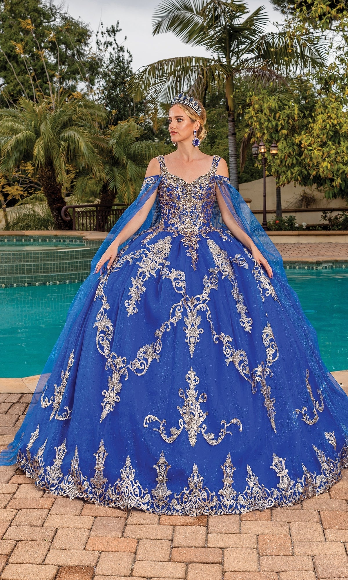A woman stands by a pool in the Quinceanera Dress 1651 By Dancing Queen, an A-line ball gown with silver embroidery and off-the-shoulder sleeves, surrounded by trees and greenery.